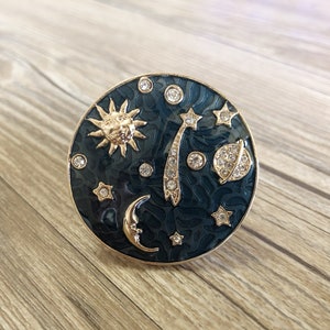 May include: A round, dark blue trinket box with a gold rim and a celestial design. The box features a gold sun, moon, stars, and a comet with rhinestones.