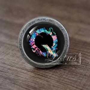 May include: A round, silver-coloured cabinet knob with a black background and a colourful abstract design. The design features swirls of pink, blue, green, and yellow paint.