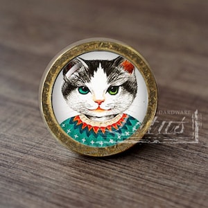 Może przedstawiać: Okrągły, mosiężny gałka do szafki z wkładką szklaną przedstawiającą kreskówkową kotkę w turkusowym i pomarańczowym swetrze. Kotka ma białe futro z czarnymi znaczeniami i zielone oczy.