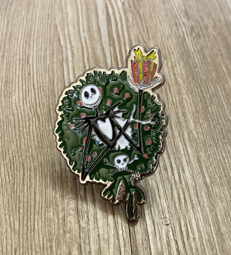 以下が含まれることがあります： ナイトメア・ビフォア・クリスマスのジャック・スケリントンがクリスマスツリーに扮した、黒と白のエナメルピン。赤いリボンと黄色のギフトボックスが乗っています。