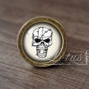 Puede incluir: Un pomo de armario redondo de color dorado antiguo con un diseño de calavera blanca. La calavera está rodeada por un diseño de alambre de púas y un fondo de lunares blancos.
