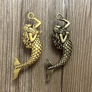 May include: Two gold-toned mermaid charms with a detailed design. The mermaids are holding a ring in their hands and have long flowing hair. The charms are perfect for adding a touch of whimsy to jewellery or crafts.