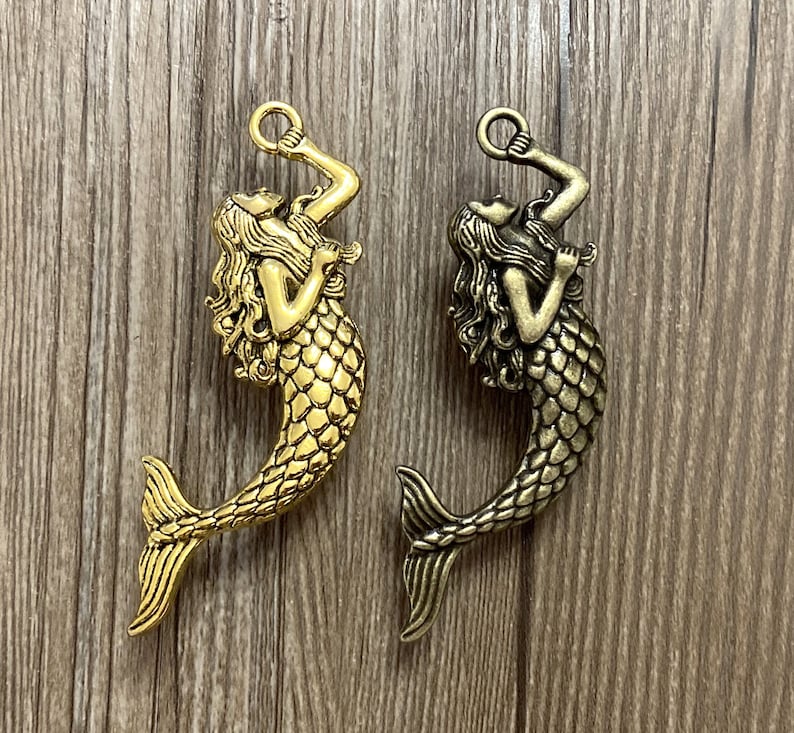 K&ouml;nnte beinhalten: Zwei goldfarbene Meerjungfrauen-Anh&auml;nger mit detailliertem Design. Die Meerjungfrauen halten einen Ring in ihren H&auml;nden und haben langes, flie&szlig;endes Haar. Die Anh&auml;nger sind perfekt, um Schmuck oder Bastelarbeiten mit einem Hauch von Fantasie zu versehen.