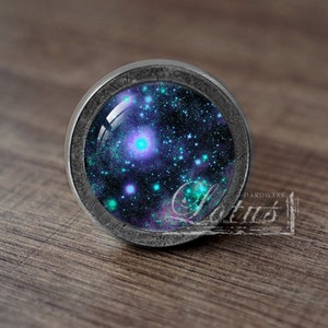 Puede incluir: Un pomo plateado con un inserto de vidrio que presenta un diseño de galaxia con estrellas azules, moradas y verdes.