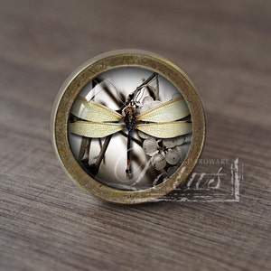 Könnte beinhalten: Ein runder, messingfarbener Schrankknopf mit einem Glaseinsatz, der eine Libelle mit gelben Flügeln auf weißem Hintergrund zeigt. Die Libelle sitzt auf einem Zweig mit weißen Beeren.