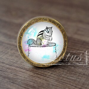 May include: A round, gold-coloured cabinet knob with a glass insert featuring a black and white illustration of a squirrel sitting on a tree stump. The background is a watercolour wash of blue, pink, and green.