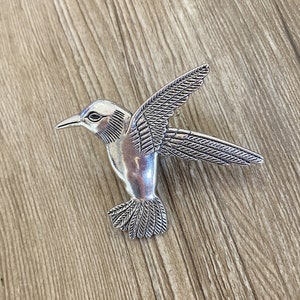 Peut inclure: Une broche en argent en forme de colibri avec des ailes détaillées et un corps texturé.