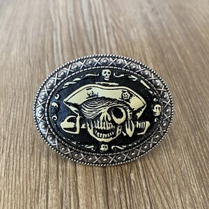以下が含まれることがあります： 海賊の帽子をかぶった白黒のドクロが描かれた銀色のベルトバックル。ドクロはクロスボーンのデザインで、バックルは装飾的な縁取りが施されています。