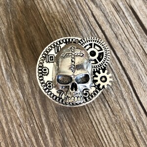 May include: Silver-toned skull charm with a cross on its forehead, surrounded by gears and a clock face with Roman numerals.