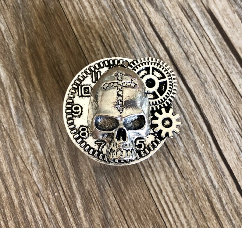 May include: Silver-toned skull charm with a cross on its forehead, surrounded by gears and a clock face with Roman numerals.