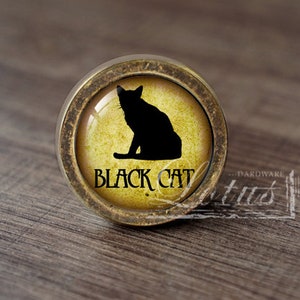 Może przedstawiać: Okrągła gałka do szafki w stylu antycznym z czarną sylwetką kota na żółtym tle. Słowa "BLACK CAT" są wydrukowane pod kotem. Gałka ma brązową metalową obwódkę.