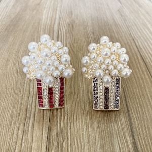 May include: Two gold-tone popcorn brooches with pearl accents. One brooch has a red and white striped popcorn box and the other has a black and white striped popcorn box.