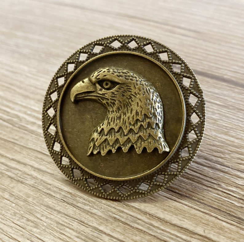 May include: A bronze-coloured metal eagle head charm with a decorative border. The eagle is facing right and has a detailed design.