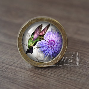 Puede incluir: Un pomo de armario redondo de color latón con un inserto de vidrio que presenta un colibrí verde flotando cerca de una flor morada.