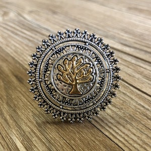 May include: A silver ring featuring a gold tree design in the centre. The ring is surrounded by a decorative silver border.