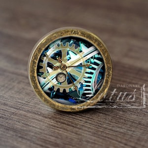 May include: A round, brass-colored knob with a glass insert featuring a steampunk design of gears and clockwork. The design is in shades of blue, green, and gold.
