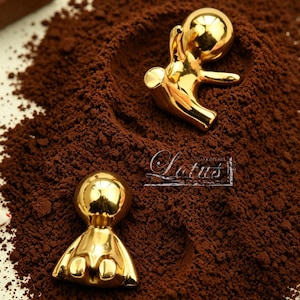 May include: Two gold-colored decorative figurines, one in a human-like form with outstretched arms and the other in a seated pose. The figurines are set against a backdrop of dark brown powder, possibly coffee grounds. The word "Lotus" is visible.