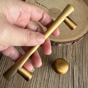 May include: A gold-toned cabinet pull and a matching knob. The pull is a long, straight bar with perpendicular cylindrical ends. The knob is a small, rounded disc. Both hardware pieces have a brushed metal finish.