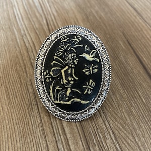 Peut inclure: Une bague en argent avec un camée ovale noir représentant un dessin détaillé d'une fée avec des ailes et un colibri. Le camée est entouré d'une bordure argentée avec un motif répété de X et de points.