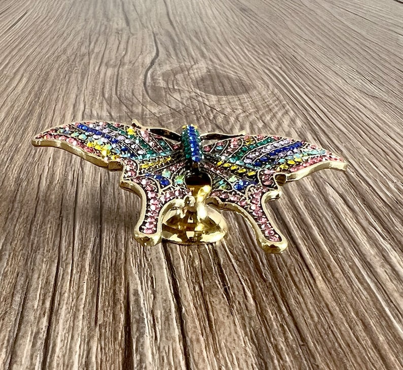 May include: A gold butterfly-shaped door knob with a colourful crystal design. The knob is mounted on a wooden surface.