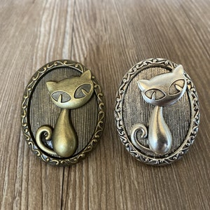 Könnte beinhalten: Zwei dekorative Broschen in Katzenform, eine in Gold und eine in Silber, mit einem detaillierten Design und einer Vintage-Ästhetik.