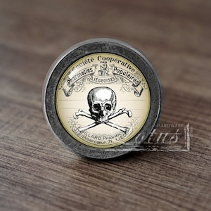 Könnte beinhalten: Ein runder, silberfarbener Schrankknopf mit einem schwarz-weißen Bild eines Totenkopfs und gekreuzter Knochen. Das Bild ist von einem gelben Etikett mit dem Text "Societe Cooperative Pharmacies Populaires Liegeoises" und "Gilllard Pharmacien 71, Liege" umgeben.