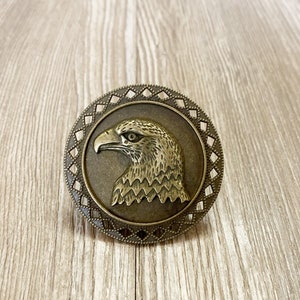 May include: A bronze-colored metal eagle head ornament with a decorative border. The eagle's head is facing left and has a detailed design.