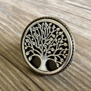 May include: A silver and bronze circular pendant featuring a detailed tree of life design. The tree has a dark background and intricate branches with leaves.
