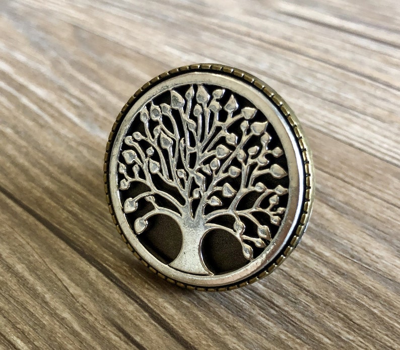 May include: A silver and bronze circular pendant featuring a detailed tree of life design. The tree has a dark background and intricate branches with leaves.