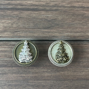 Può includere: Due ornamenti rotondi per albero di Natale color argento con un design a forma di albero di Natale in rilievo color argento. Gli ornamenti hanno un bordo perlato e uno sfondo marrone.