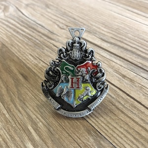 Puede incluir: Un amuleto de metal plateado con el escudo de la casa de Hogwarts con un diseño de esmalte verde, amarillo, rojo y azul. El escudo presenta una serpiente, un león, un tejón y un águila. Las palabras "Draco Dormiens Nunquam Titillandus" están inscritas alrededor del escudo.