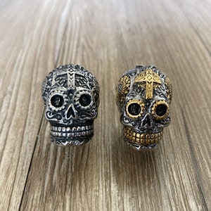 May include: Two silver skull rings with gold accents. The skulls have intricate details and a cross on the forehead.