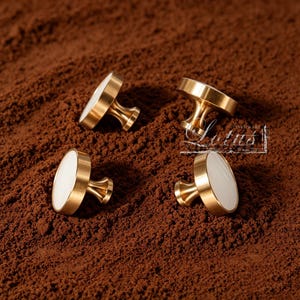 May include: Four gold cabinet knobs with white oval inlays are arranged on a brown surface. The knobs have a classic design with a rounded base and a decorative stem. The knobs are likely for furniture or cabinetry.