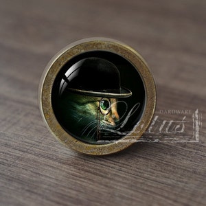 Op de afbeelding: Een ronde, messingkleurige kastknop met een glazen inzetstuk met een kat die een zwarte bolhoed en een bril draagt. De kat heeft groene ogen en een witte snor.