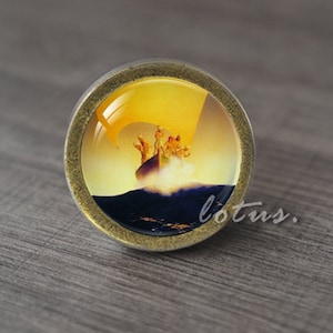 May include: A round, brass-colored cabinet knob with a glass insert featuring a vintage illustration of a boat with people in it, sailing on a yellow and blue ocean.
