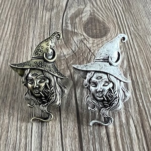 May include: Two silver tone witch charm pendants with a third eye and a crescent moon on the hat. The witch is wearing a pointed hat and has long hair.