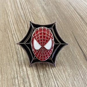 Może przedstawiać: Czarna i srebrna szpilka emaliowana z czerwoną i czarną maską Spider-Mana z białymi oczami. Maska jest umieszczona na czarnym tle z pajęczyną.