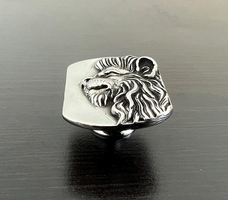 Pode incluir: Um bot&atilde;o de arm&aacute;rio de cabe&ccedil;a de le&atilde;o de prata com um design detalhado. O bot&atilde;o &eacute; montado em uma base quadrada.