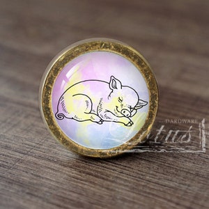 以下が含まれることがあります： 水彩画風の寝ている豚のデザインが施されたガラス製のインサートが付いた、丸い金色のキャビネットノブ。豚は黒で縁取られており、目を閉じて横向きに寝ています。