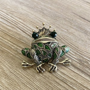May include: A gold-tone frog brooch with green enamel and green rhinestone eyes. The frog is wearing a gold-tone crown.