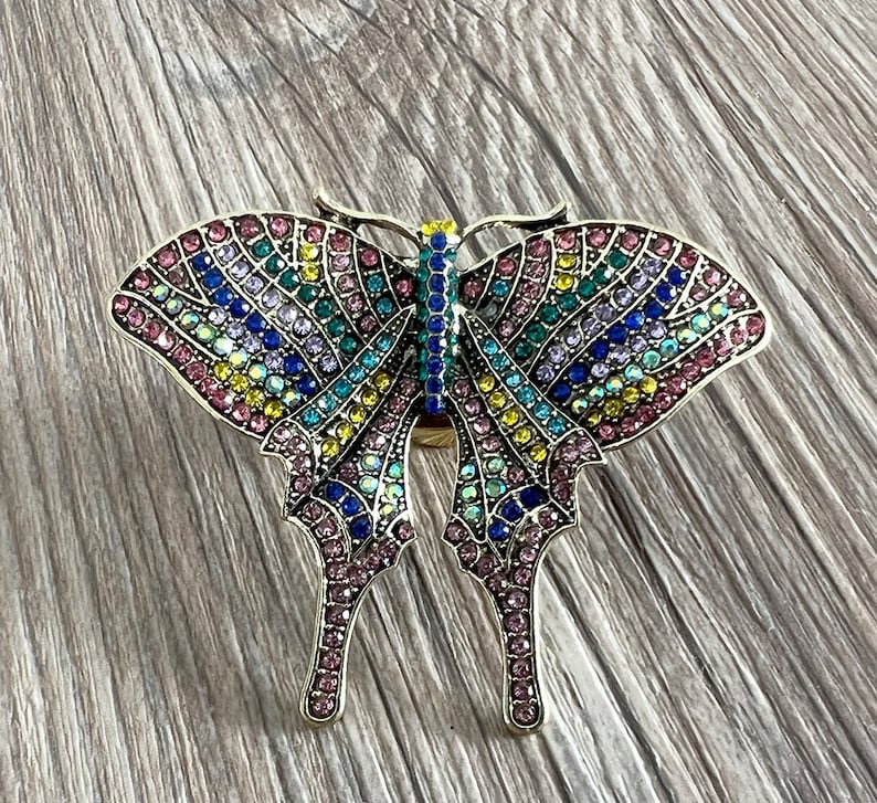 May include: A colourful butterfly brooch with pink, blue, green and yellow rhinestones. The butterfly is made of metal and has a detailed design.