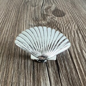 May include: A silver seashell-shaped decorative object with a detailed, textured surface. The seashell is on a wooden background.