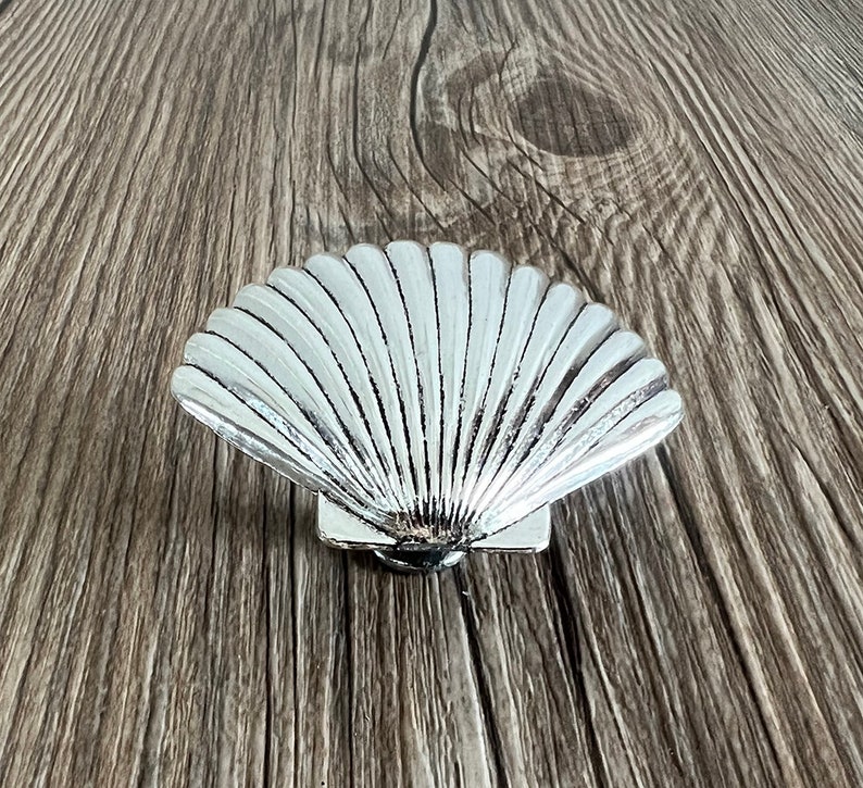 May include: A silver seashell-shaped decorative object with a detailed, textured surface. The seashell is on a wooden background.