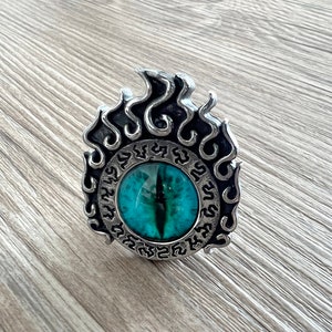 以下が含まれることがあります： 炎のデザインとルーンの円に囲まれた、中央に青い目を持つ銀製のペンダント