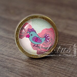 Può includere: Un pomello per armadio rotondo in ottone con un inserto in vetro che presenta un uccello dei cartoni animati con accenti rosa e viola, circondato da rose rosa su uno sfondo bianco.