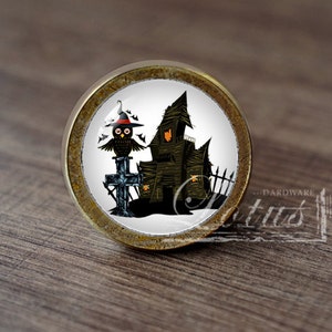 Pode incluir: Um botão de armário redondo de cor latão com um fundo branco e um design temático de Halloween. O design apresenta uma coruja preta usando um chapéu de bruxa empoleirada em uma cruz, e uma casa assustadora verde escura com uma cerca no fundo.