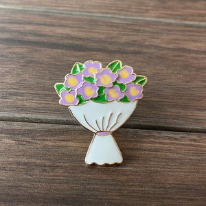 May include: A gold-toned enamel pin depicting a cartoon bouquet of purple and yellow flowers with green leaves. The flowers are arranged in a white paper wrapper with a gold outline.