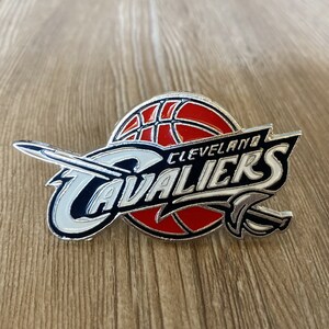 May include: A silver and black Cleveland Cavaliers pin with a red basketball and a sword design. The pin features the team's name and city.