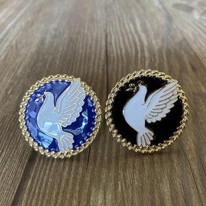 May include: Two round pins with gold rims, each featuring a white dove design. One pin has a blue background and the other has a black background.