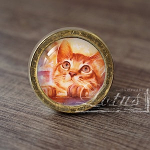 May include: A round, gold-colored cabinet knob with a glass insert featuring a painting of a ginger kitten looking up. The kitten has green eyes and is sitting on a purple surface.